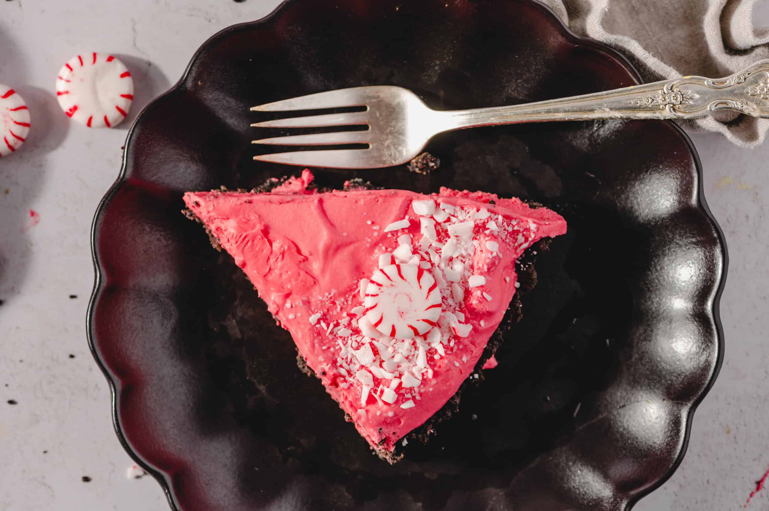 Delicious peppermint chocolate cheesecake slice with pink frosting, peppermint candy, and white chocolate shavings on a black decorative plate. Perfect for holiday desserts and festive occasions.