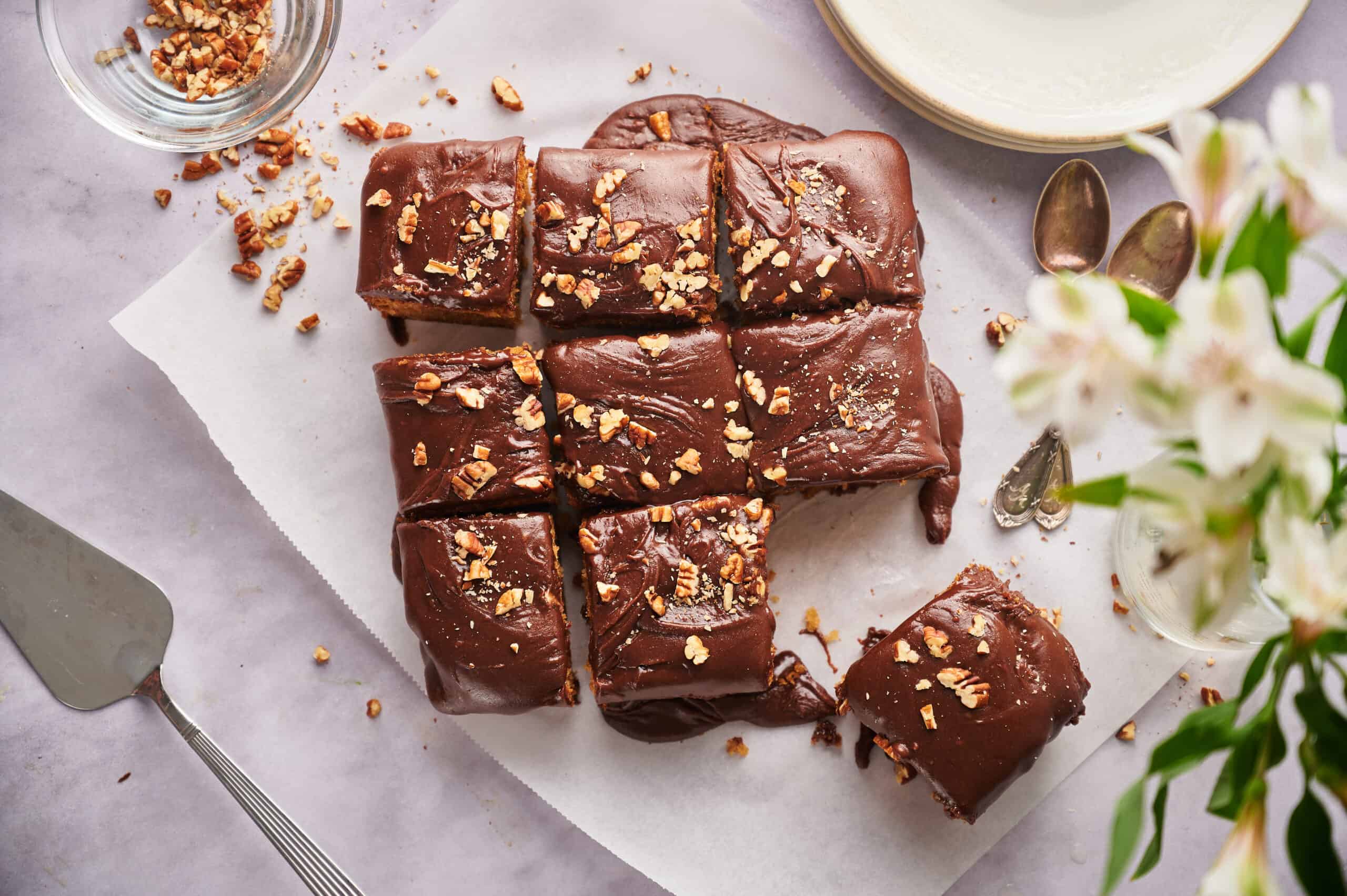 Rich chocolate fudge squares topped with chopped nuts on parchment paper with dessert utensils and flowers in the scene.