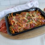 Delicious pepperoni and sausage baked pasta in a black tray, topped with cheese and fresh herbs, served with side of sliced pepperoni, marinara sauce, and chopped parsley on a wooden platter.