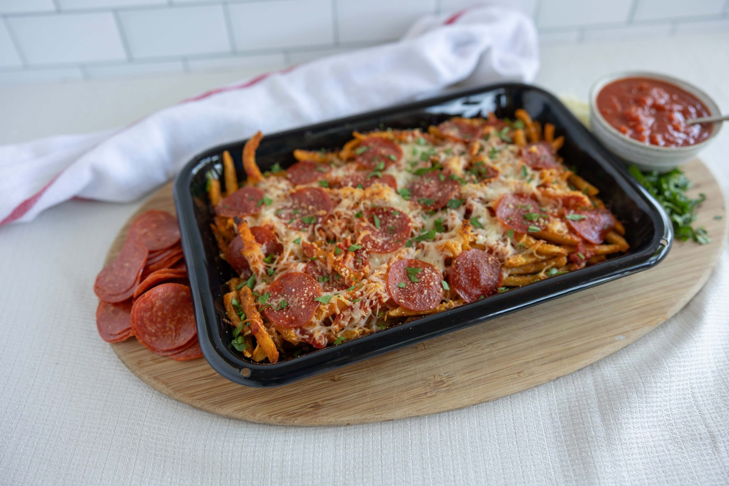 Delicious pepperoni and sausage baked pasta in a black tray, topped with cheese and fresh herbs, served with side of sliced pepperoni, marinara sauce, and chopped parsley on a wooden platter.