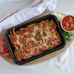 Slice pepperoni pizza with melted cheese, spaghetti, and tomato sauce served on a black tray with fresh herbs and shredded cheese on a wooden board.