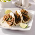 Shredded beef tacos with lime wedges on white plate, mexican street food, flavorful pulled beef in soft tacos, garnished with cilantro, served with fresh lime slices, appetizing dinner or lunch dish, Hey Recipe image.