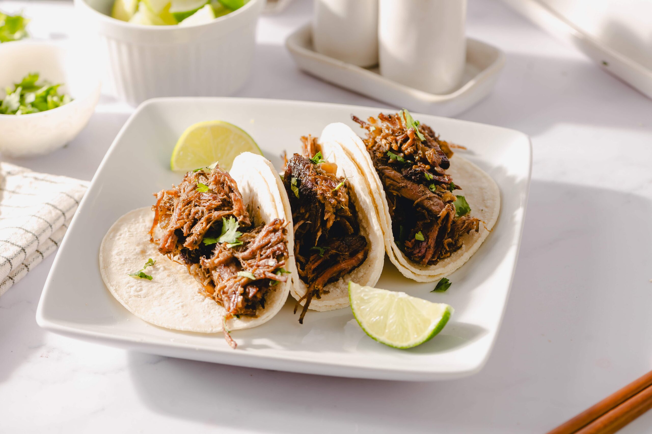 Shredded beef tacos with lime wedges on white plate, mexican street food, flavorful pulled beef in soft tacos, garnished with cilantro, served with fresh lime slices, appetizing dinner or lunch dish, Hey Recipe image.