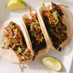 Shredded beef tacos with lime wedges on white plate, Mexican cuisine, savory street food, delicious homemade taco filling, flavorful beef recipe, garnished with cilantro, perfect for dinner or lunch.