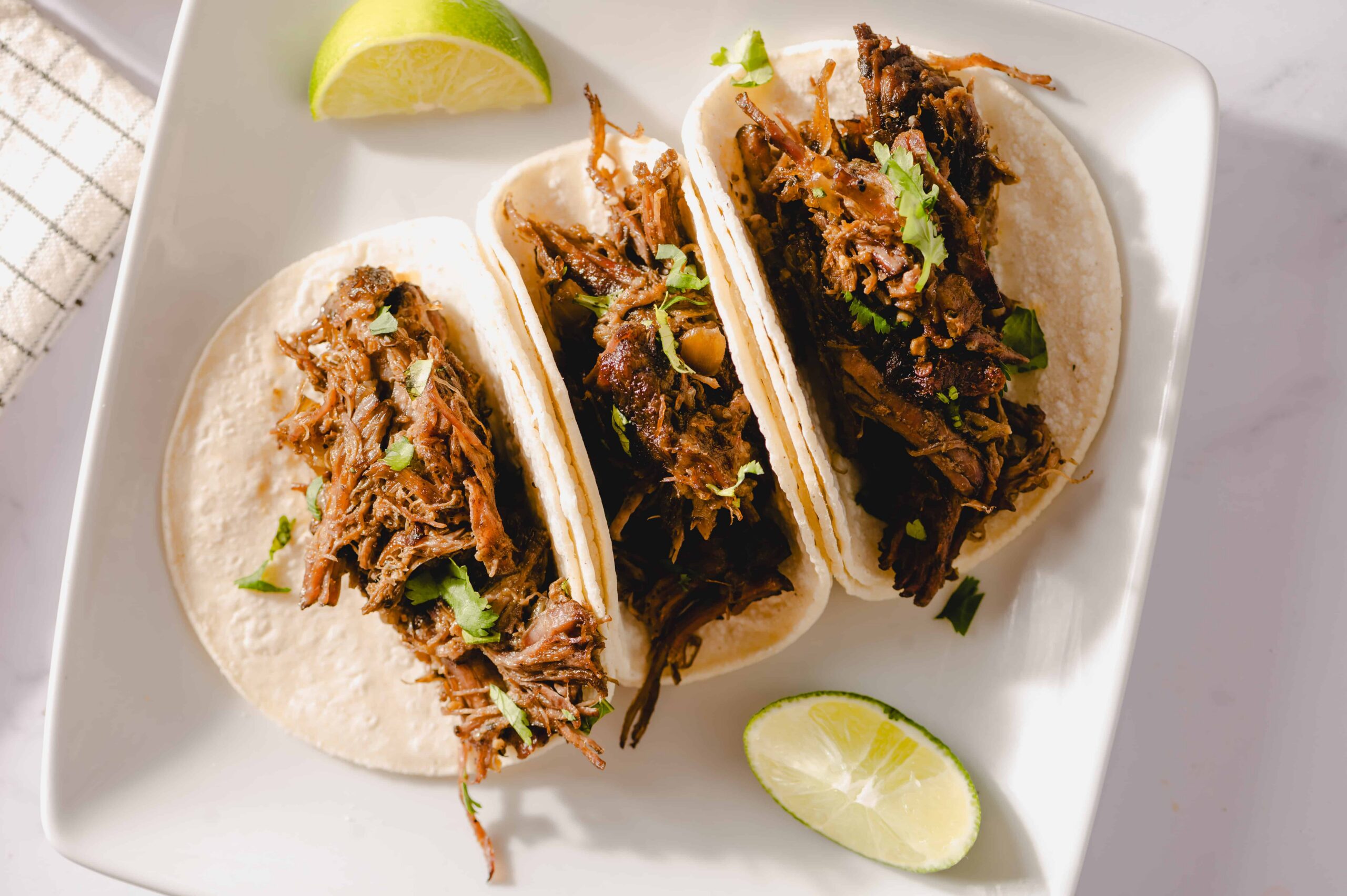 Shredded beef tacos with lime wedges on white plate, Mexican cuisine, savory street food, delicious homemade taco filling, flavorful beef recipe, garnished with cilantro, perfect for dinner or lunch.