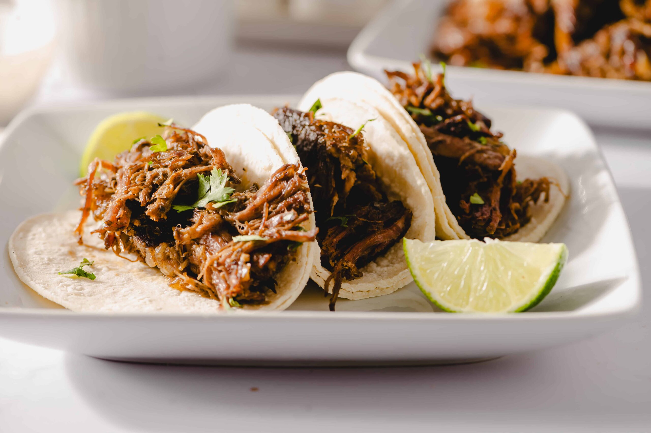 Tender shredded beef tacos on soft corn tortillas garnished with fresh cilantro and lemon wedges, served on a white rectangular plate for an authentic Mexican cuisine experience.