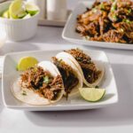 Slow cooker shredded beef tacos with lime wedges on a white plate, featuring tender beef in soft tortillas, garnished with cilantro, served with side bowls of fresh lime and chopped green onions, delectable Mexican street food.