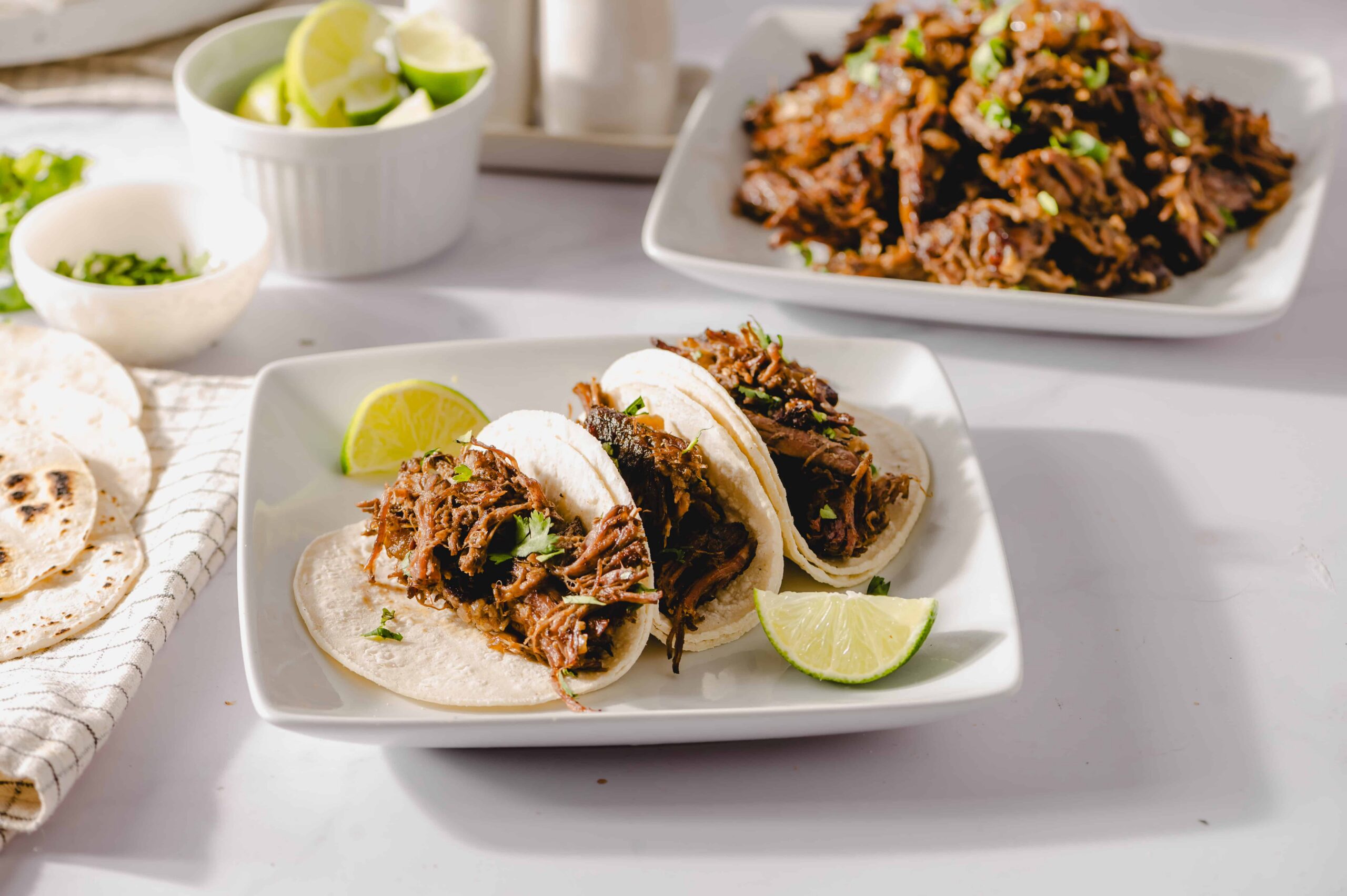 Slow cooker shredded beef tacos with lime wedges on a white plate, featuring tender beef in soft tortillas, garnished with cilantro, served with side bowls of fresh lime and chopped green onions, delectable Mexican street food.