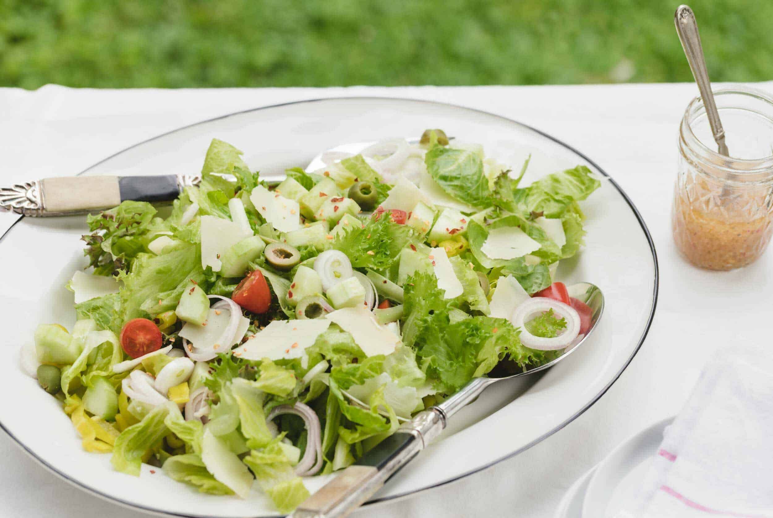 Fresh garden salad with mixed greens, cherry tomatoes, cucumbers, olives, and cheese, served on a white plate with a dressing jar on the side.