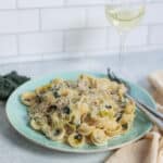 Creamy chicken and mushroom tortellini pasta dish served on a light turquoise plate with shredded cheese, fresh herbs, and a fork; white wine in a glass, Parmesan cheese wedge, and black truffle on a kitchen countertop.