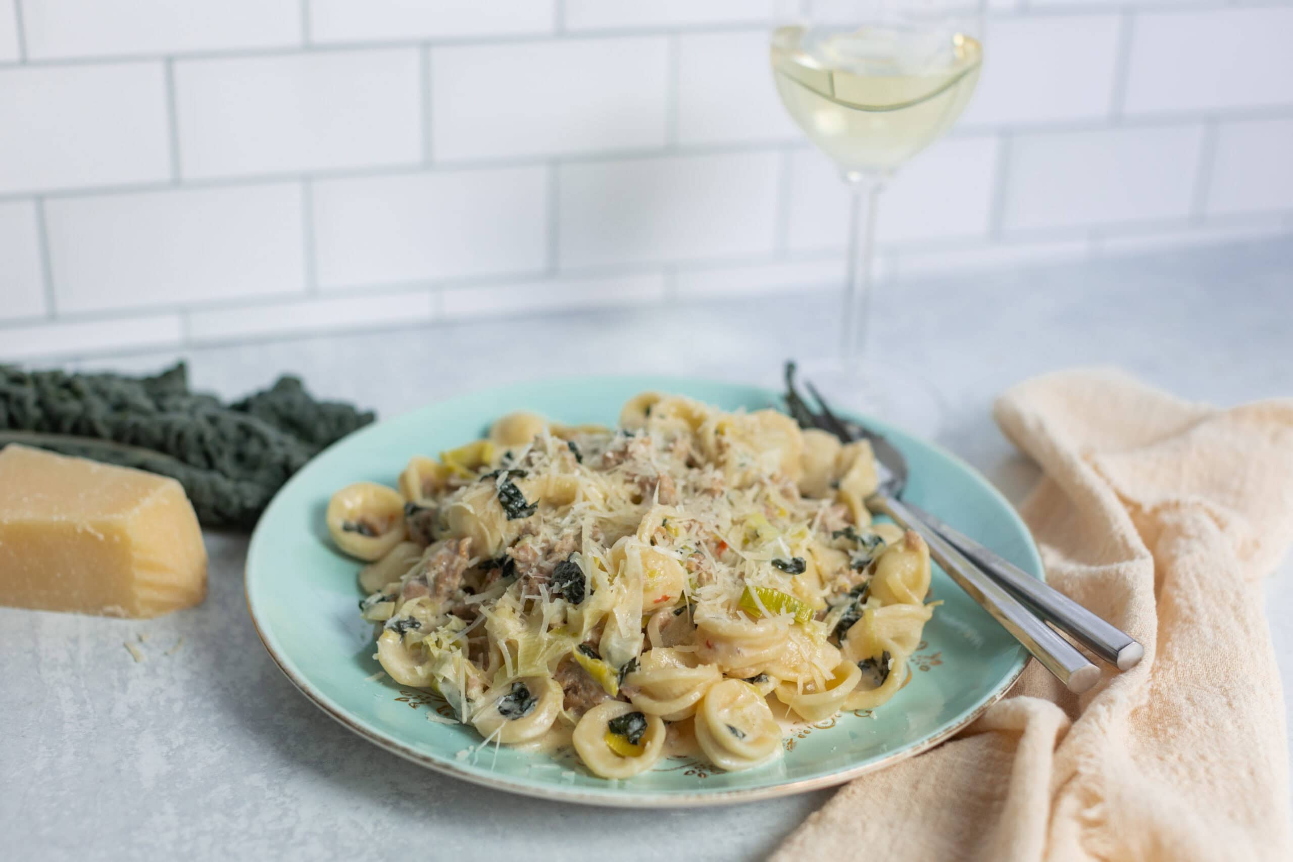 Creamy chicken and mushroom tortellini pasta dish served on a light turquoise plate with shredded cheese, fresh herbs, and a fork; white wine in a glass, Parmesan cheese wedge, and black truffle on a kitchen countertop.