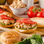 Fresh chicken salad sandwiches on sandwich buns with sliced tomatoes and lettuce, served on a rustic wooden board with fresh tomatoes and bread rolls, perfect for a quick lunch or picnic.