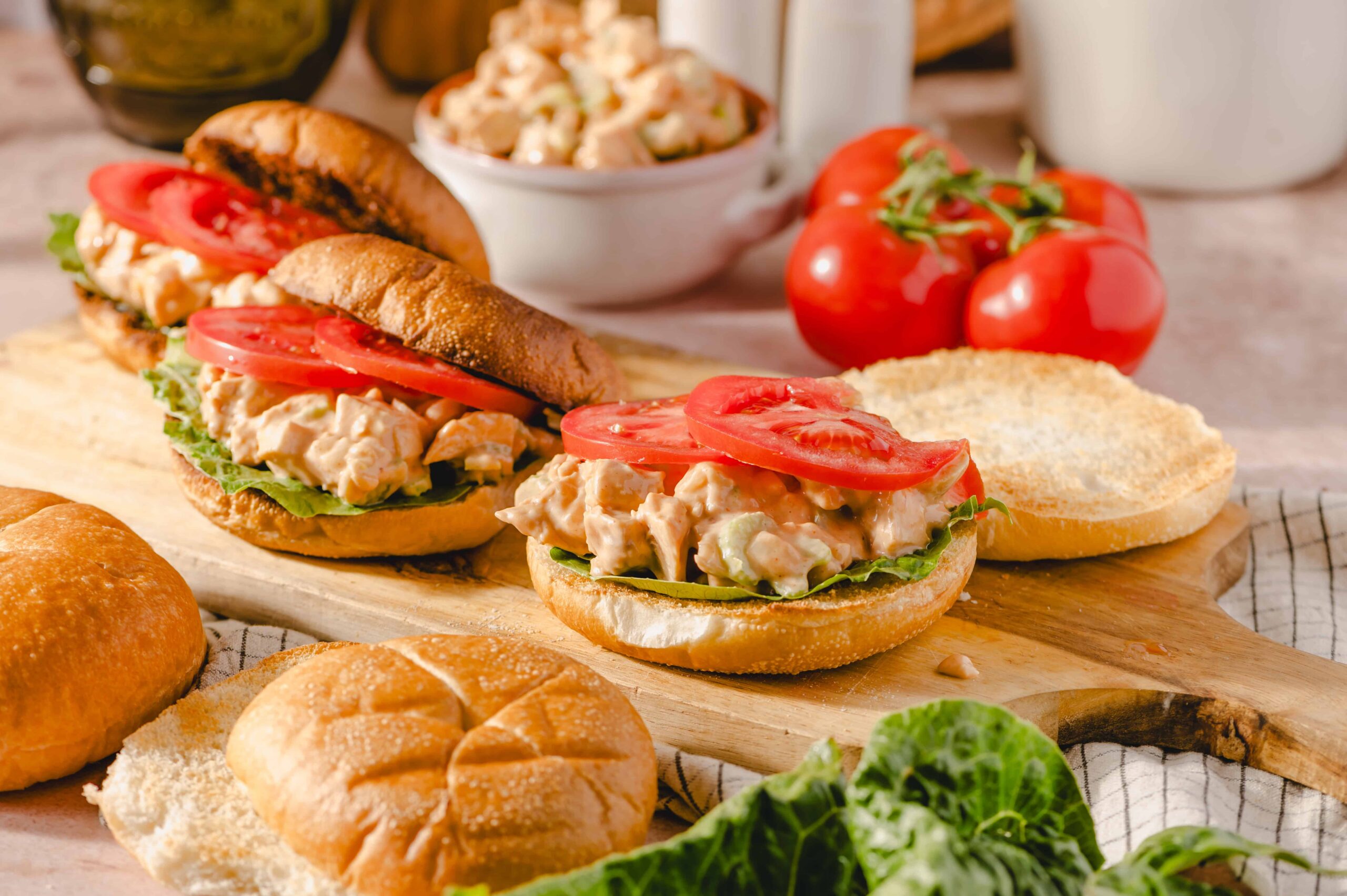 Fresh chicken salad sandwiches on sandwich buns with sliced tomatoes and lettuce, served on a rustic wooden board with fresh tomatoes and bread rolls, perfect for a quick lunch or picnic.
