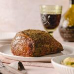 Juicy roasted beef tenderloin seasoned with herbs and spices on a white plate, served with a glass of red wine and a bottle of balsamic vinegar in the background. Perfect for dinner.