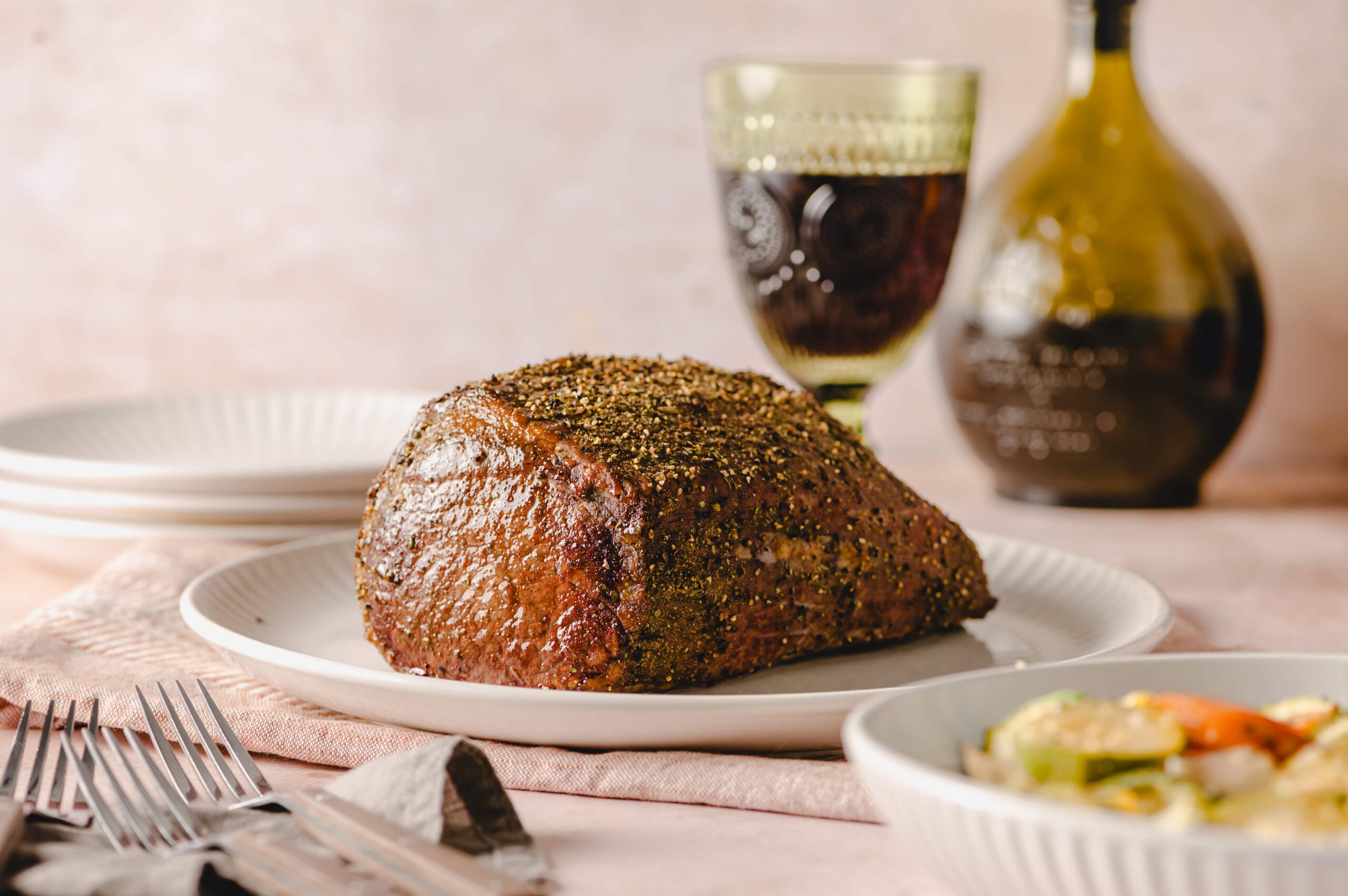Juicy roasted beef tenderloin seasoned with herbs and spices on a white plate, served with a glass of red wine and a bottle of balsamic vinegar in the background. Perfect for dinner.