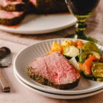 Juicy, perfectly cooked prime rib steak coated with flavorful herb crust served with roasted zucchini, carrots, and potatoes on a white ceramic plate for a delicious and hearty meal.