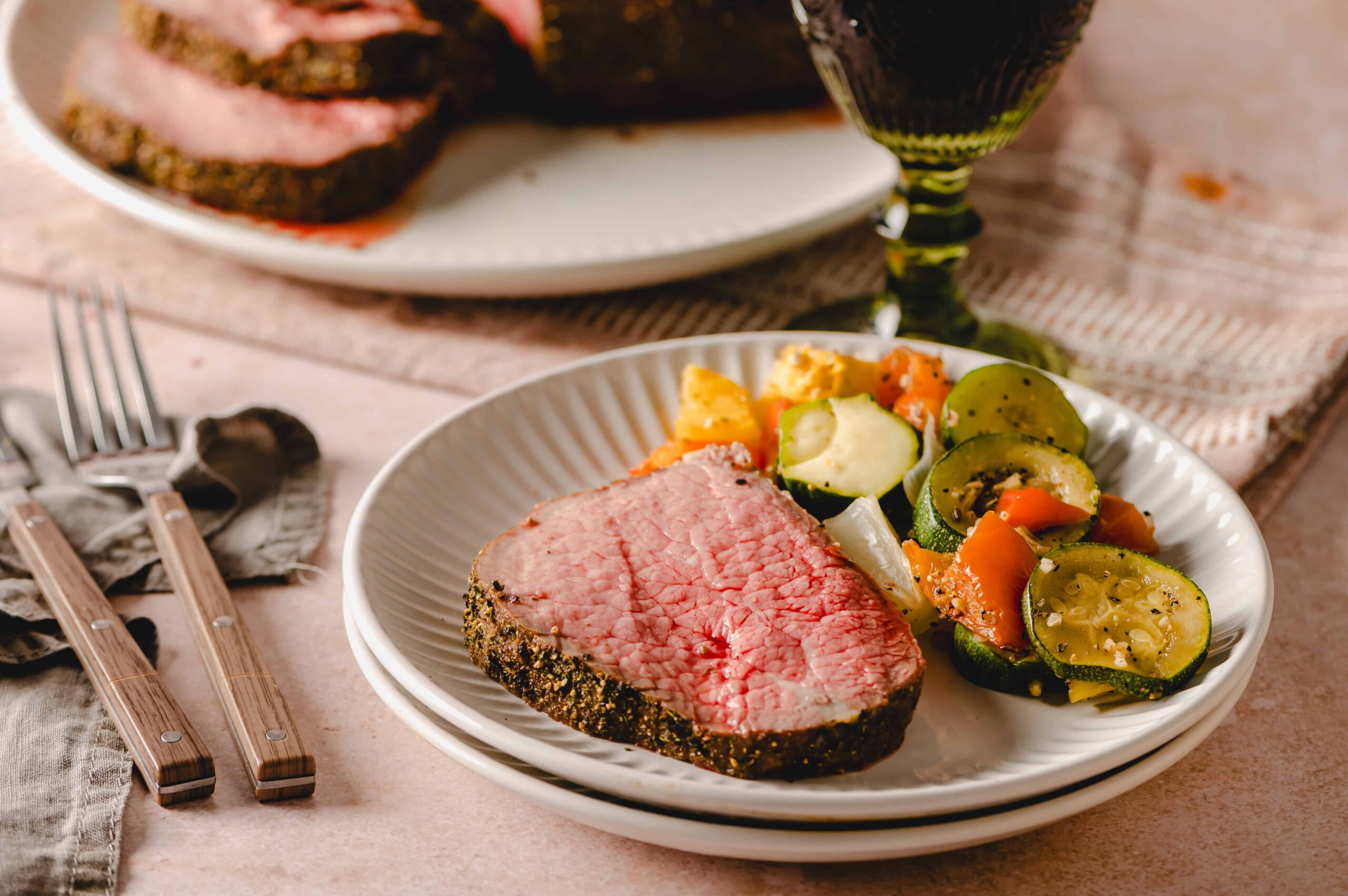 Juicy, perfectly cooked prime rib steak coated with flavorful herb crust served with roasted zucchini, carrots, and potatoes on a white ceramic plate for a delicious and hearty meal.
