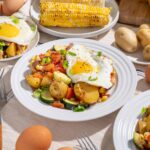 Creamy potato hash with vegetables topped with fried eggs, served on white plates, surrounded by fresh potatoes, eggs, and corn on a rustic table setting.