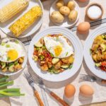 Soft-boiled eggs served with sautéed vegetables and fried eggs on white plates, surrounded by fresh corn, potatoes, eggs, and seasoning on a rustic wooden table.