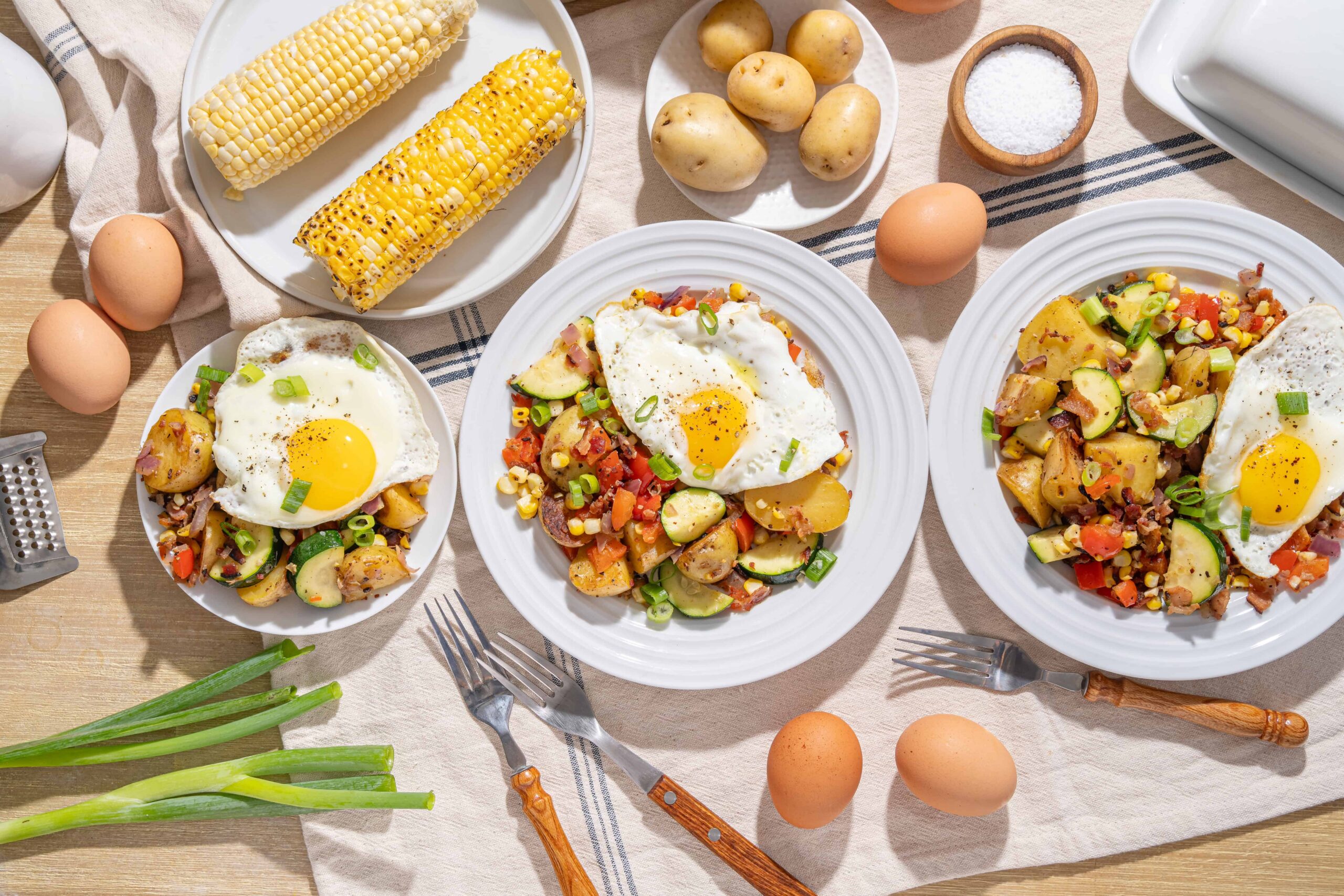 Soft-boiled eggs served with sautéed vegetables and fried eggs on white plates, surrounded by fresh corn, potatoes, eggs, and seasoning on a rustic wooden table.