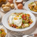 Fried egg served over roasted vegetables and hash in a white plate, with side dishes of corn on the cob, boiled potatoes, and eggs on a rustic table setting.