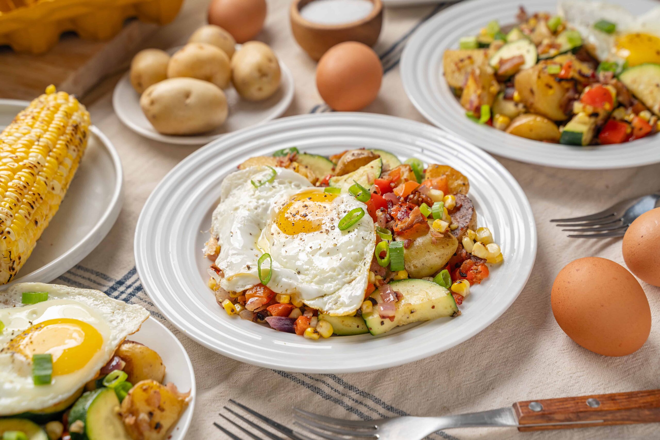 Fried egg served over roasted vegetables and hash in a white plate, with side dishes of corn on the cob, boiled potatoes, and eggs on a rustic table setting.