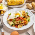Roasted potato and vegetable breakfast hash topped with sunny side up eggs, garnished with chopped green onions, served on white plates with fresh ingredients in the background.