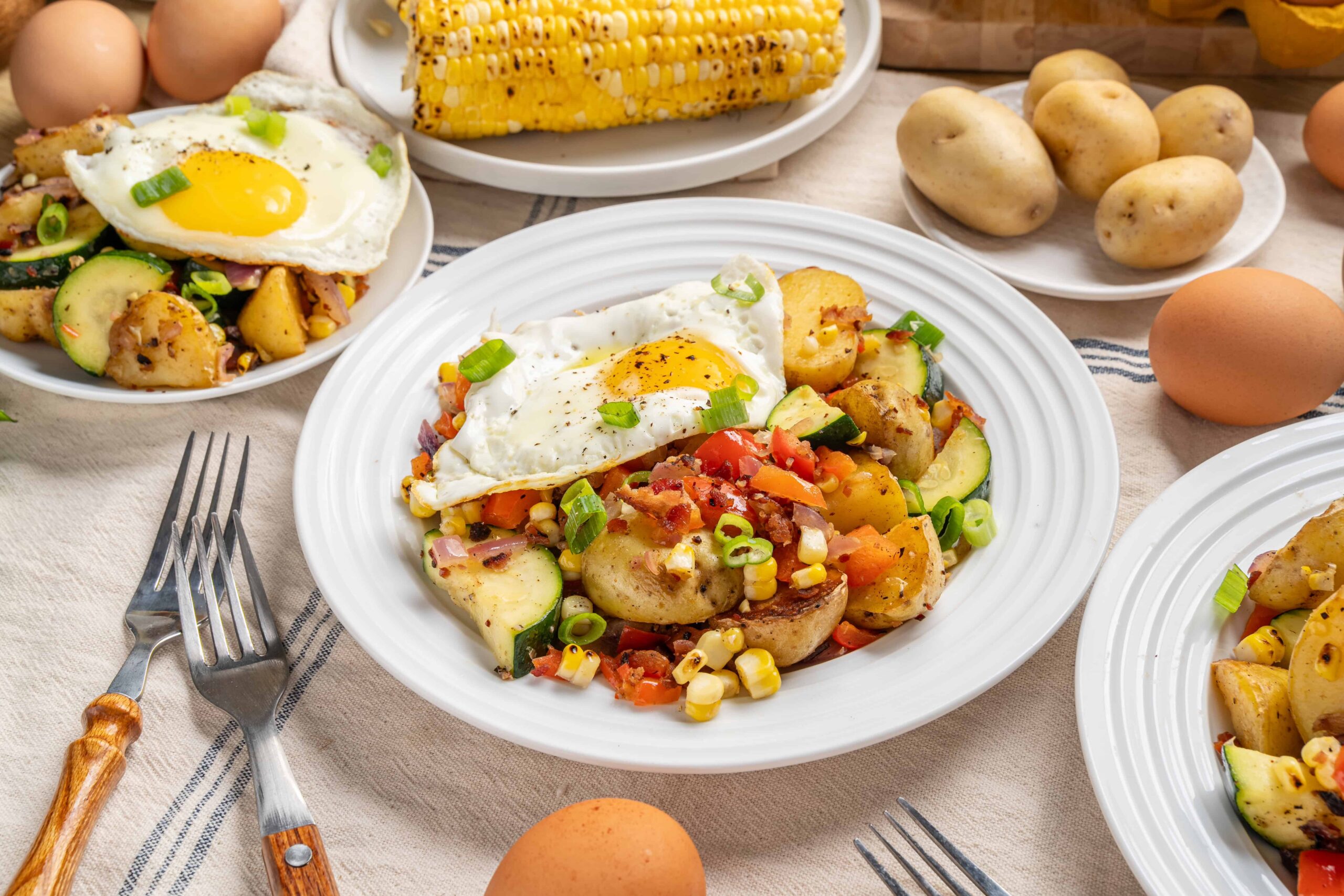 Roasted potato and vegetable breakfast hash topped with sunny side up eggs, garnished with chopped green onions, served on white plates with fresh ingredients in the background.