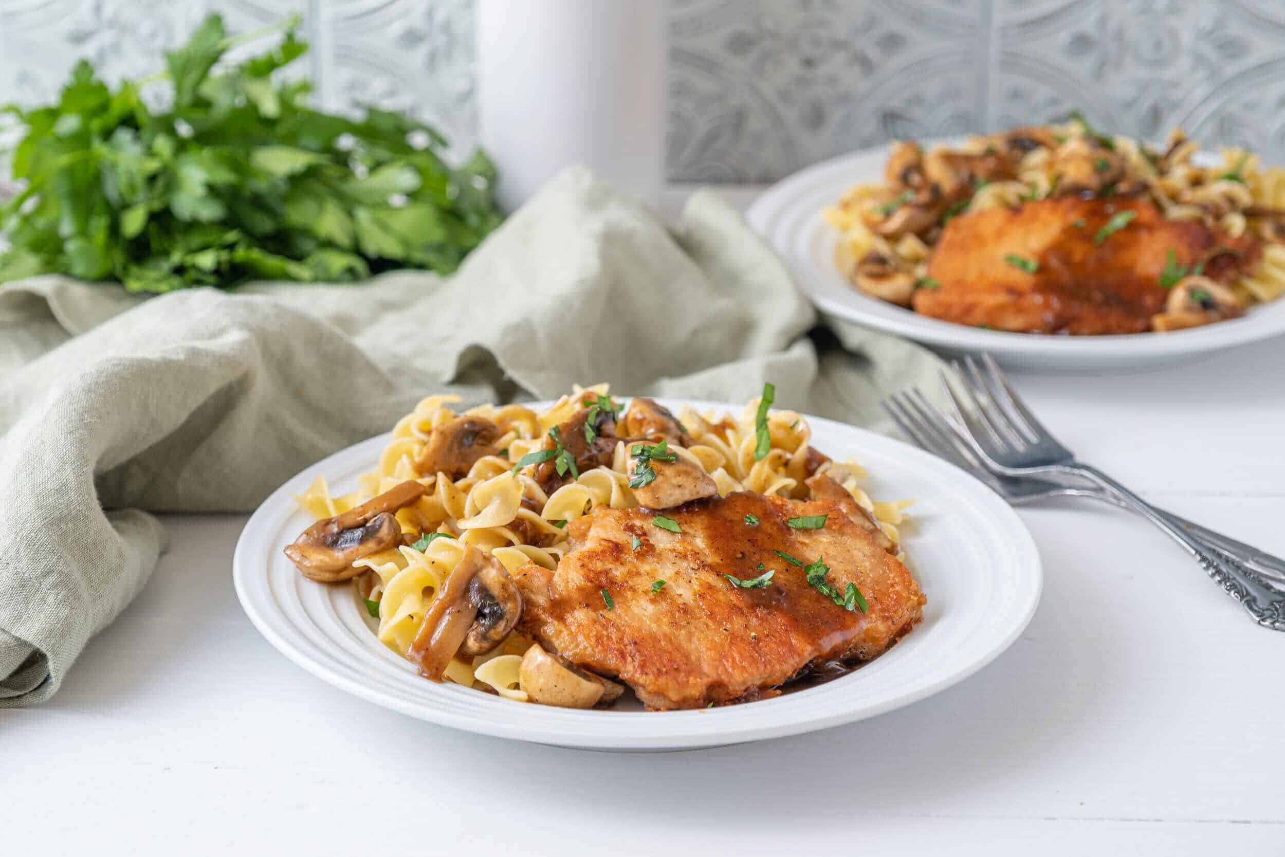 Juicy pan-seared chicken breast with mushroom pasta on a white plate, garnished with fresh herbs, served with a side of green leafy vegetables on a casual dining table.