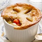 Golden-brown chicken pot pie in a ramekin topped with flaky pastry, served on a blue and white patterned plate with a silver spoon, highlighting comforting homemade classic American cuisine.