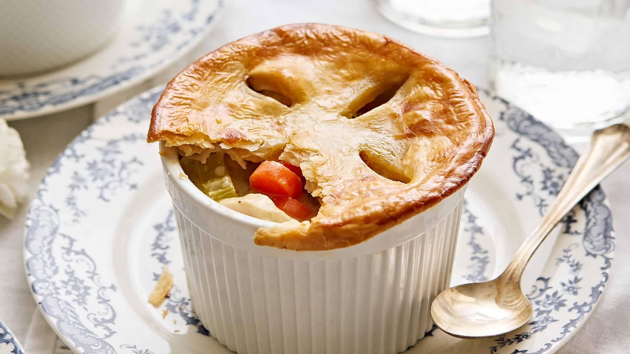Golden-brown chicken pot pie in a ramekin topped with flaky pastry, served on a blue and white patterned plate with a silver spoon, highlighting comforting homemade classic American cuisine.
