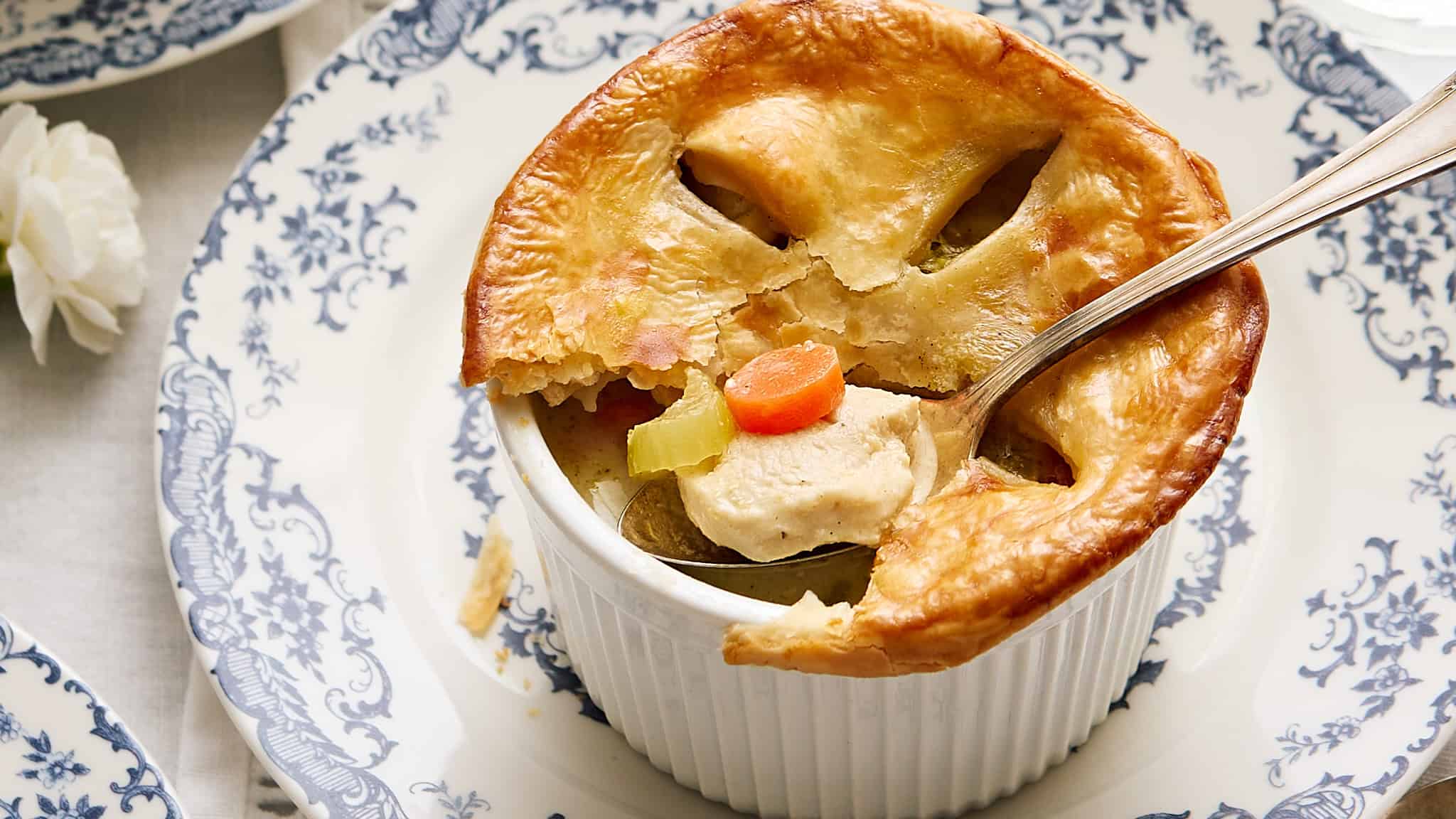 Creamy chicken pot pie with golden flaky crust, served in a white ramekin on a blue and white patterned plate. Comes with tender chicken, carrots, and celery in a savory broth. Perfect comfort food.