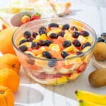 Fresh fruit salad with citrus, grapes, banana, and strawberries in a glass bowl on a white surface; vibrant and healthy summertime dessert.