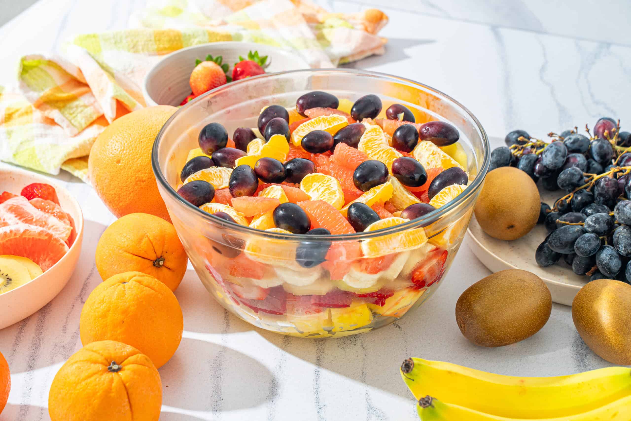 Fresh fruit salad with citrus, grapes, banana, and strawberries in a glass bowl on a white surface; vibrant and healthy summertime dessert.