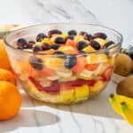 Fresh fruit salad with mixed berries, oranges, strawberries, bananas, and kiwis in a glass bowl on a white marble surface.