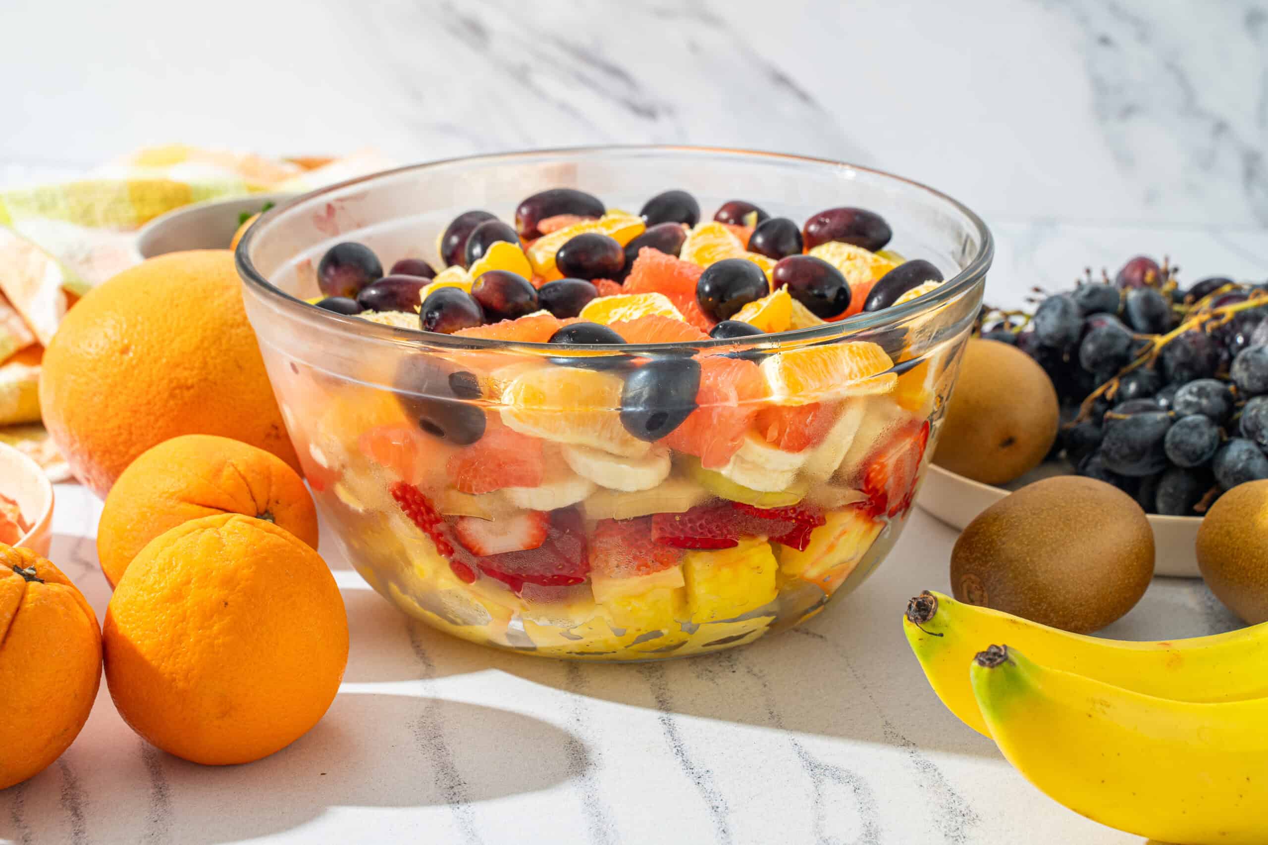 Fresh fruit salad with mixed berries, oranges, strawberries, bananas, and kiwis in a glass bowl on a white marble surface.