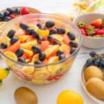 Assorted fresh fruit salad with oranges, strawberries, grapes, kiwis, and bananas in a clear glass bowl on a white surface.