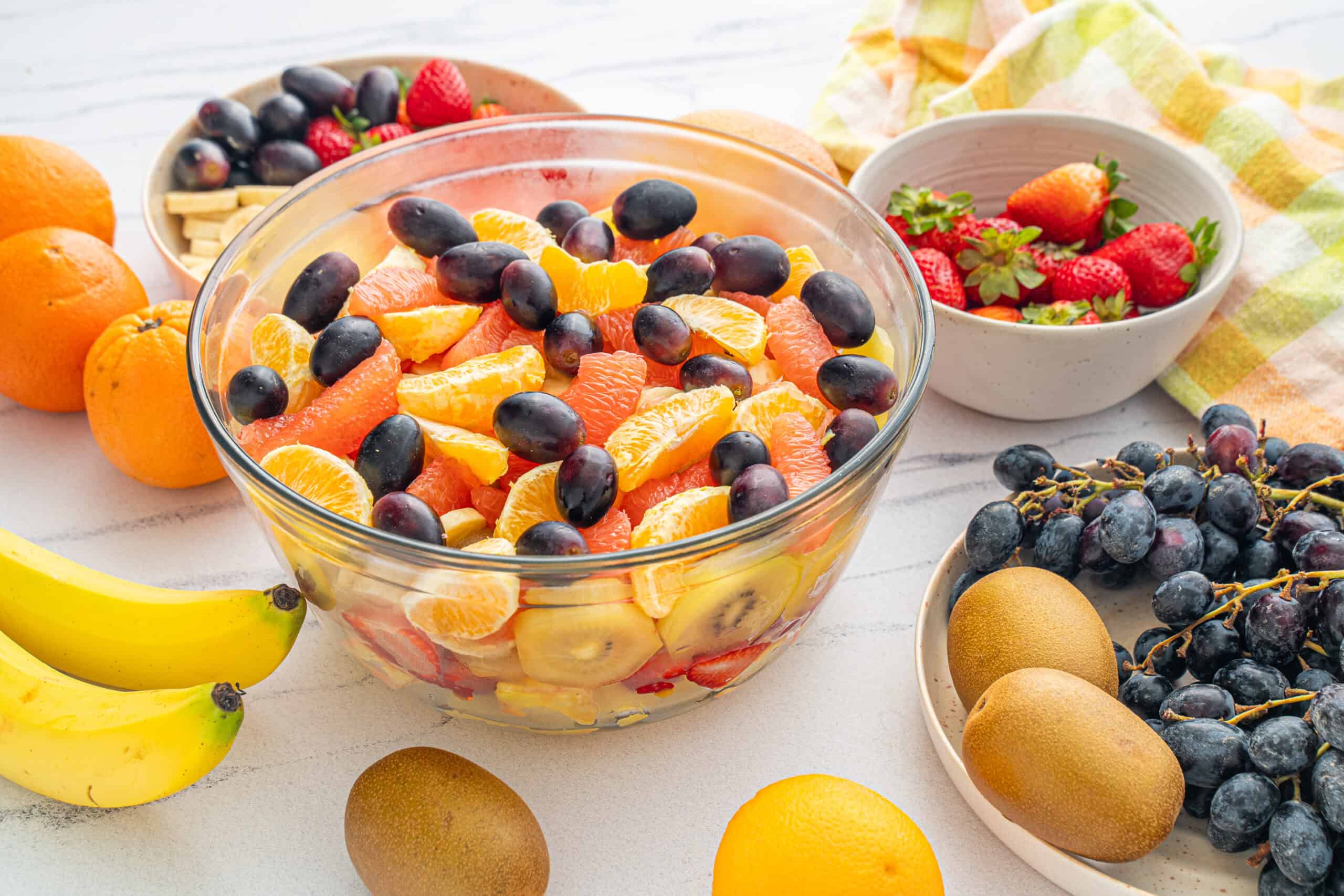 Assorted fresh fruit salad with oranges, strawberries, grapes, kiwis, and bananas in a clear glass bowl on a white surface.