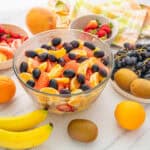 Juicy mixed fruit salad with oranges, bananas, grapes, strawberries, and kiwi, served in a clear glass bowl on a white countertop with fresh whole fruits around.