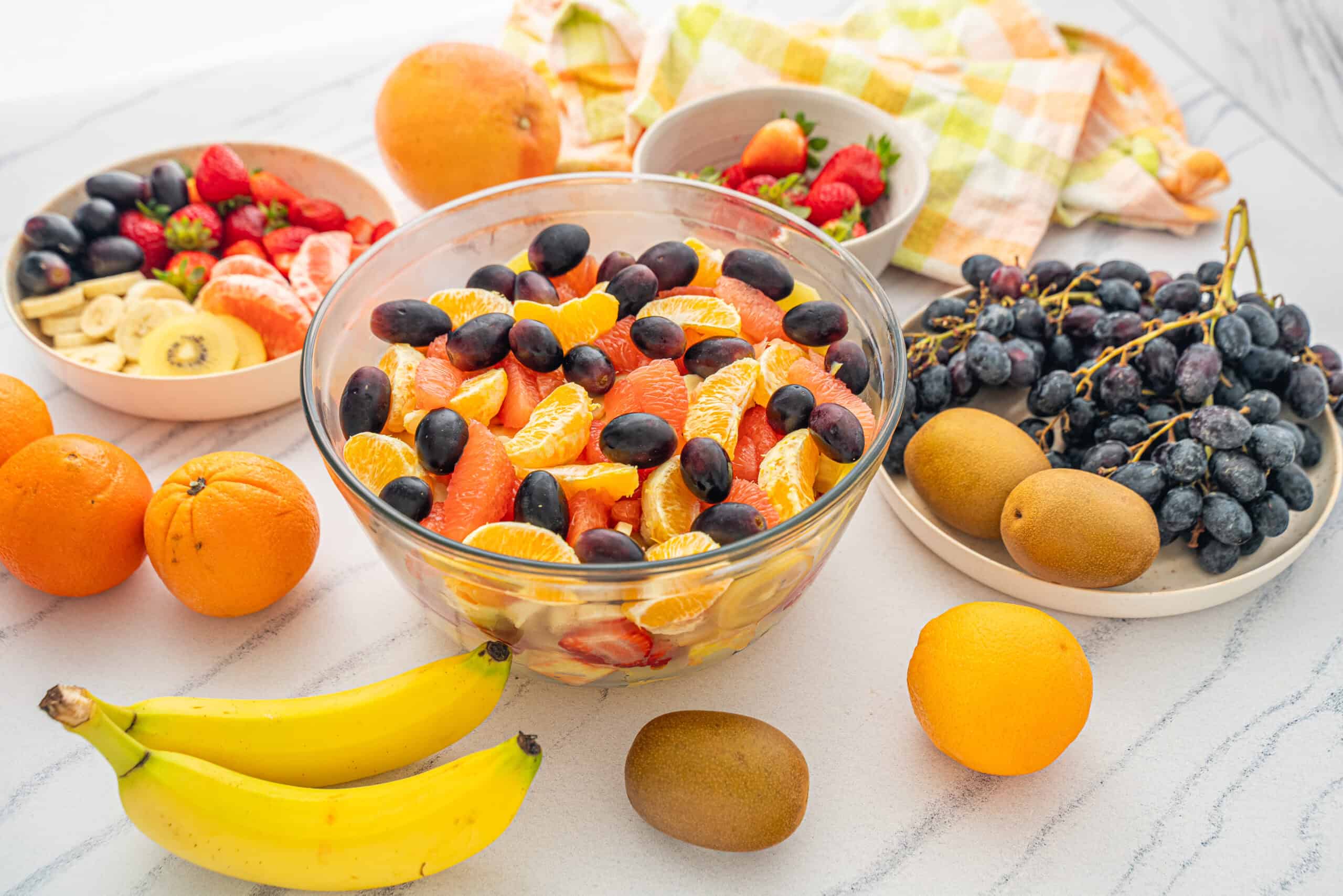 Juicy mixed fruit salad with oranges, bananas, grapes, strawberries, and kiwi, served in a clear glass bowl on a white countertop with fresh whole fruits around.
