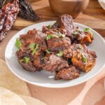 Slow-cooked beef barbacoa with tender, flavorful meat garnished with fresh cilantro, served on a white plate with chili peppers and traditional Mexican ingredients in the background.