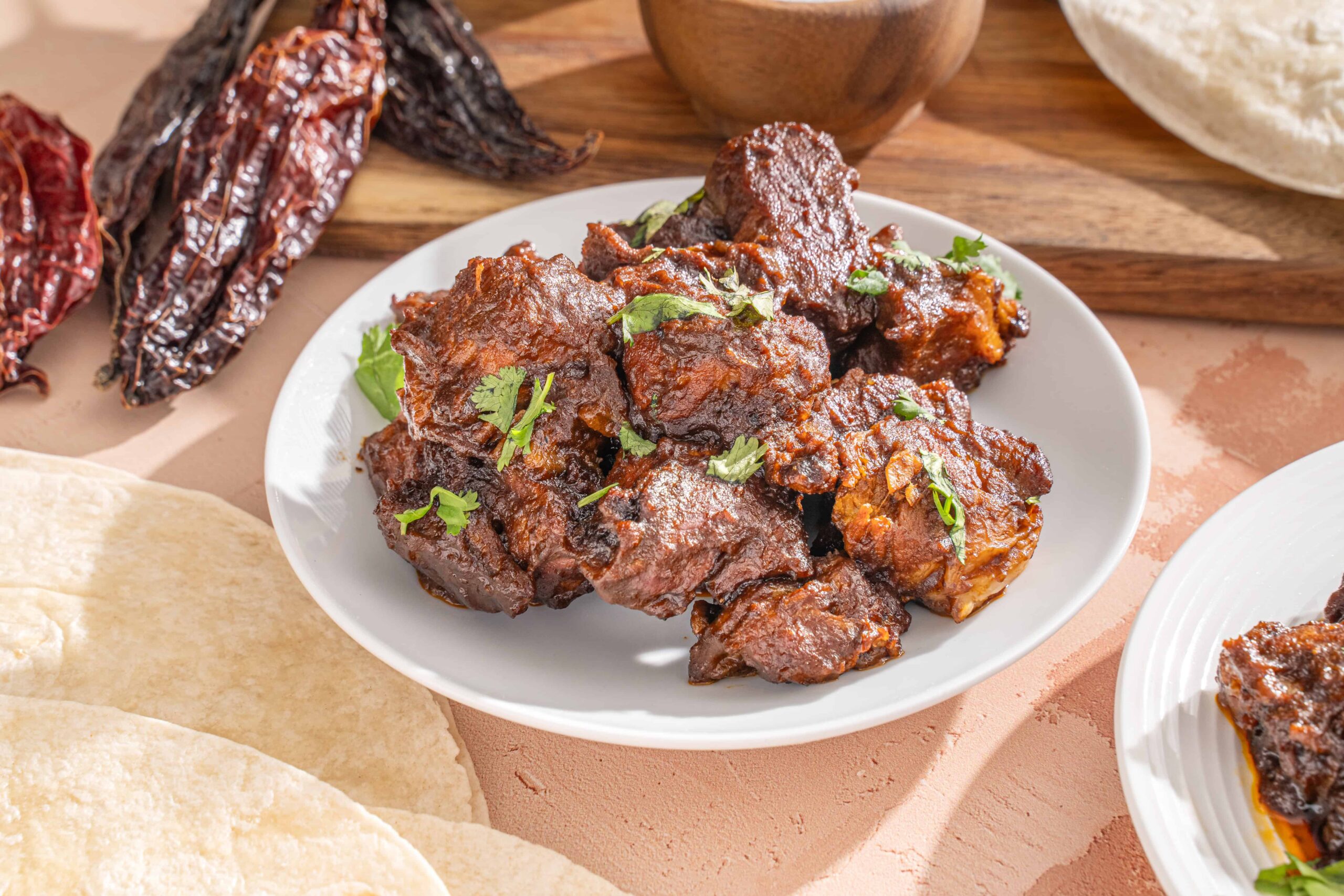 Slow-cooked beef barbacoa with tender, flavorful meat garnished with fresh cilantro, served on a white plate with chili peppers and traditional Mexican ingredients in the background.