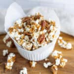 Crunchy caramel pecan popcorn in a white paper-lined bowl on a wooden cutting board, perfect for sweet snacks or movie nights.