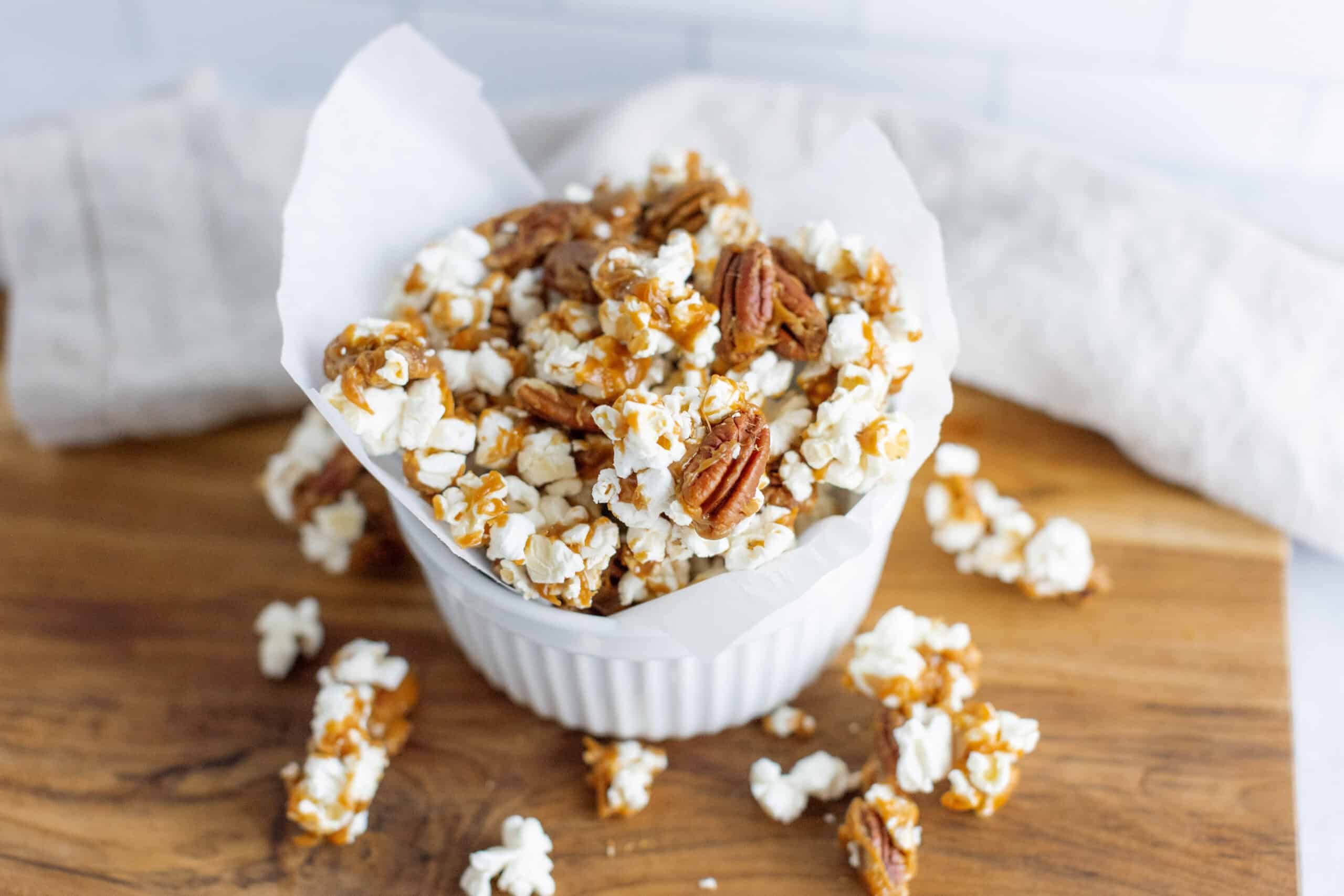 Crunchy caramel pecan popcorn in a white paper-lined bowl on a wooden cutting board, perfect for sweet snacks or movie nights.