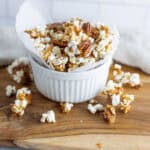 Sweet caramel popcorn with pecans in a white bowl on a wooden cutting board.