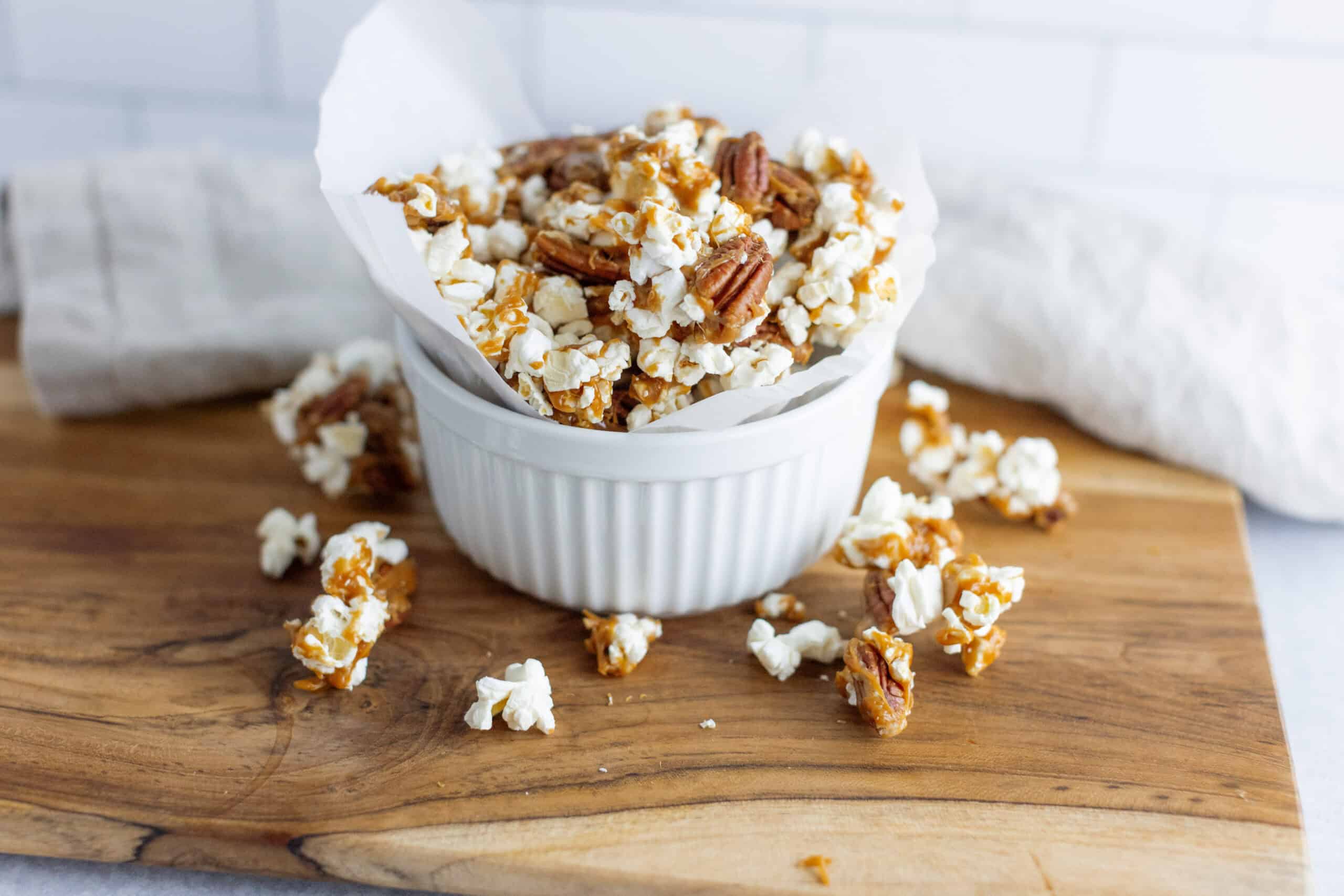 Sweet caramel popcorn with pecans in a white bowl on a wooden cutting board.