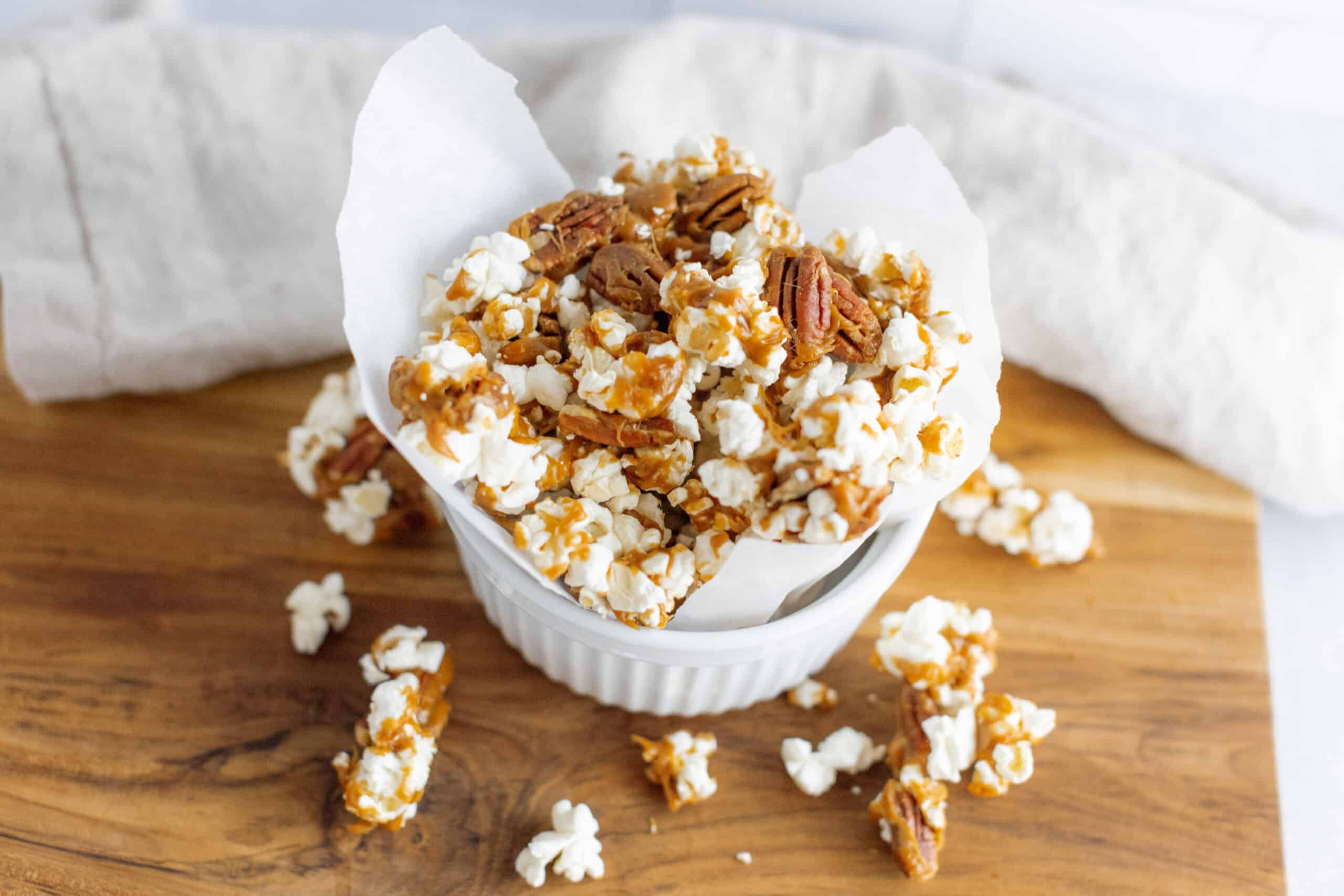 Sweet caramel popcorn with pecans in a white ramekin on a wooden surface, perfect for snack or party treat.