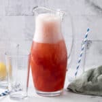 Refreshing strawberry lemonade in a glass pitcher with matching striped straws, served alongside two glasses, fresh lemons, and limes on a white surface with a gray cloth and marble background, perfect for summer drinks.