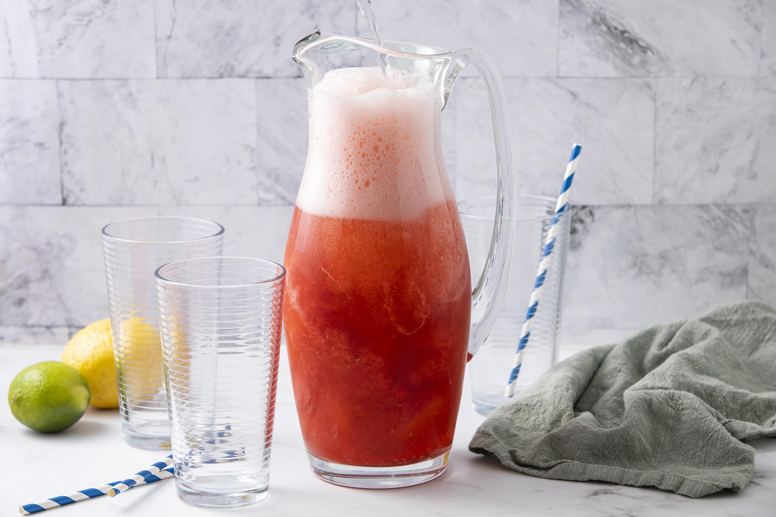 Refreshing strawberry lemonade in a glass pitcher with matching striped straws, served alongside two glasses, fresh lemons, and limes on a white surface with a gray cloth and marble background, perfect for summer drinks.