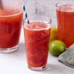 Refreshing watermelon lime juice served in tall glasses with blue and white striped straws, surrounded by fresh limes and a lemon, on a white marble surface with a rustic gray background.